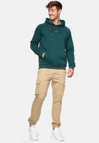 Grøn hættebluse med kængurulomme, parret med beige cargo-bukser med flere lommer. Hvide sneakers fuldender outfittet.