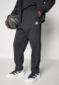 Schwarze Jogginghosen mit einem elastischen Bund, ausgestattet mit einem kleinen weißen Adidas-Logo. Kombiniert mit schwarz-weißen Sportschuhen und einem Basketball.