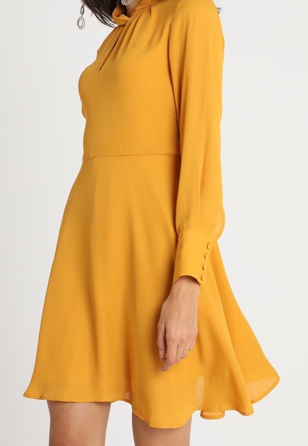 Femme portant une robe jaune moutarde à manches longues avec des poignets à boutons et une jupe évasée, debout devant un fond uni clair.