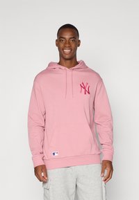 Haut à capuche rose avec une poche avant, présentant un logo brodé rose foncé sur la poitrine et un écusson MLB sur l'ourlet. Tissu doux, coupe décontractée.