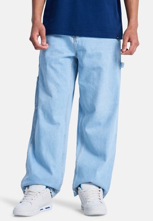 Persoon die lichtblauwe wijde spijkerbroeken en witte sneakers draagt, staand met handen ontspannen langs het lichaam tegen een effen achtergrond.