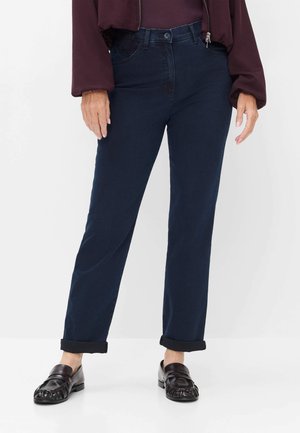 Person iført mørkeblå manchetjeans, bordeaux langærmet top og mørkebrune loafers, der står på en hvid gulv mod en hvid baggrund.