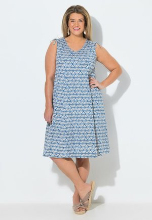 Kvinde i ærmeløs blå og hvid mønstret knælængde kjole, beige slip-on sandaler, smilende med den ene hånd på hoften mod hvid baggrund.