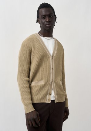 Junger Mann trägt eine beige gerippte Strickjacke mit Knöpfen über einem weißen Hemd und dunklen Hosen und steht vor einem schlichten hellen Hintergrund.