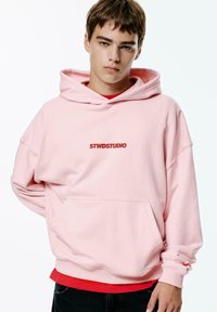 Pink hooded sweatshirt with a front pocket and "STWDSTUDIO" logo. Features loose fit, ribbed cuffs, and layered red shirt underneath.