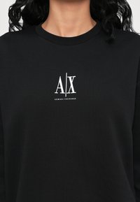 Juoda medvilninė jaunatviška palaidinė su baltu spausdintu logotipu "A/X" ir "ARMANI EXCHANGE" apačioje. Apvali iškirptė ir laisvas kirpimas.