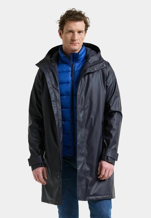 Man draagt een zwarte capuchonregenjas over een blauw gewatteerd jack en blauwe spijkerbroek, staand tegen een effen lichte achtergrond.