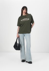 Oliivroheline ülepaisutatud T-särk, millel on valge "Barbour International" tekst, koos helesiniste flared teksade ja musta käsipagasi kotiga.