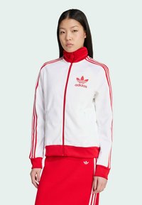 Bijela jakna sa zipom i crvenim detaljima, sa tri crvene pruge na rukavima i Adidas logom. Rebrasti manžeti i donji rub. Glatka struktura tkanine.