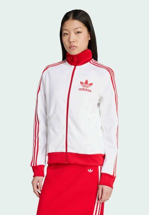 Bijela jakna sa zipom i crvenim detaljima, sa tri crvene pruge na rukavima i Adidas logom. Rebrasti manžeti i donji rub. Glatka struktura tkanine.