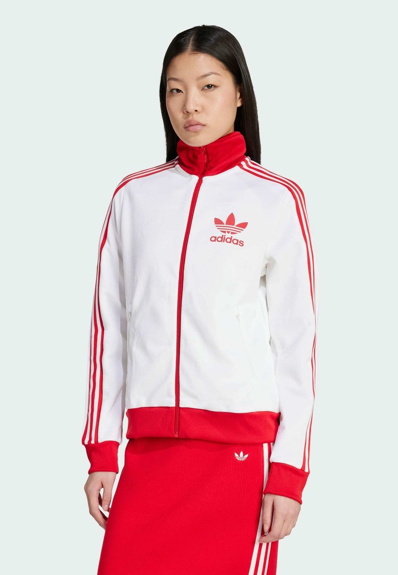 Bijela jakna sa zipom i crvenim detaljima, sa tri crvene pruge na rukavima i Adidas logom. Rebrasti manžeti i donji rub. Glatka struktura tkanine.