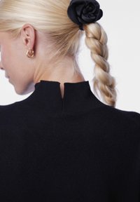 Juodas megztinis su aukštu kaklu, smulkia nugarine įpjova, minkšta tekstūra ir prisiglaudusia forma, demonstruojamas su šviesiais nupintais plaukais.