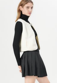 Weißer Fleece-Weste mit abgerundetem Saum, getragen über einem schwarzen Rollkragenpullover, kombiniert mit einem plissierten schwarzen Rock, der verschiedene Texturen und Formen hervorhebt.