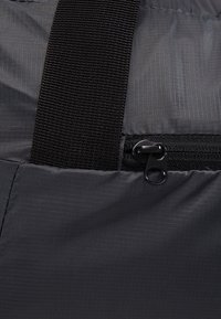 Mustan vetoketjun osittain auki tummanharmaassa ripstop-kankaassa, johon on kiinnitetty leveä musta kudottu hihna sen ylle.