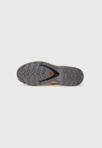 Semelle de chaussure avec un motif de bande de roulement en caoutchouc noir robuste, des crampons à profil bas, un accent rouge et une semelle intercalaire beige pour la stabilité.
