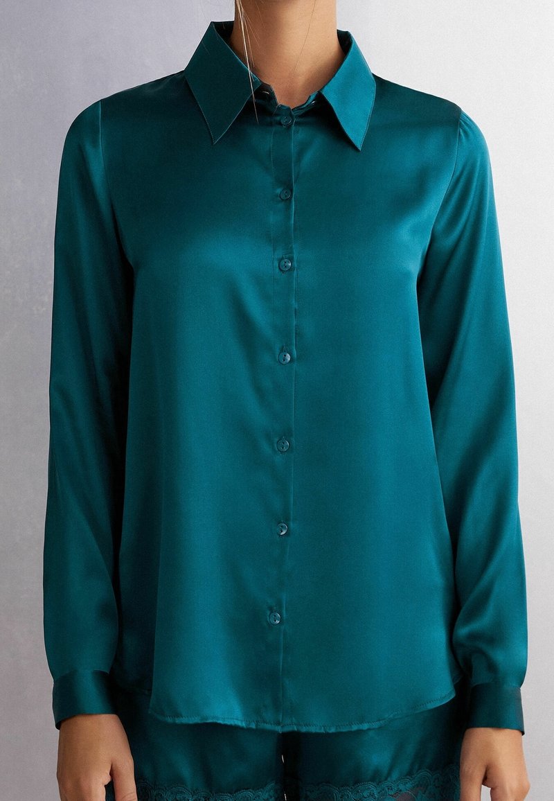 Chemise longue en satin bleu sarcelle à manches longues avec boutons et col pointu, portée sur un short assorti bordé de dentelle.
