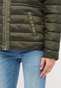 Doudoune vert olive avec couture horizontale, fermeture éclair et poche latérale. Associée à un jean en denim bleu clair, mettant en valeur une texture lisse.
