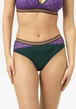Vrouw draagt donkergroene hoge tailleslip met paarse kanten rand en een gestreepte elastische tailleband, staand tegen een effen achtergrond.