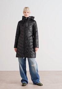 Svart quilted boblejakke med hette, med glidelåslukking, chevron-mønster og en mid-lengde design. Båret over lyseblå ripped jeans.