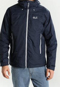 Marinblå vattentät jacka tillverkad av mjukt tyg, med en framdragkedja, två sidofickor och en huva. Logotyp på vänster bröst.