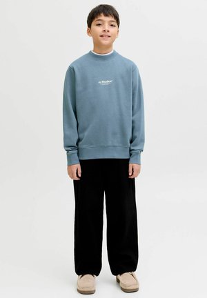 Vaaleansininen collegepaita pehmeästä kankaasta, jossa on korkea kaulus ja valkoinen logo. Yhdistetty väljien mustien housujen ja beigejen liukumiskenkien kanssa.