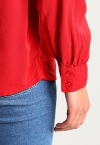 Poignet d'un chemisier rouge à manches longues avec deux boutons, porté avec un jean en denim bleu, montrant un bras et une main détendus sur un fond clair.