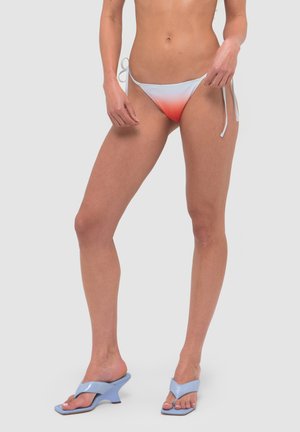 Kobieta w białe i czerwone ombre majtki bikini z wiązaniami po bokach oraz w jasnoniebieskie sandały na obcasie na jednolitym tle.