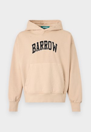 Mladý muž s kučeravými vlasmi nosiaci béžovú mikinu s kapucňou "BARROW" a zodpovedajúce šortky, biele ponožky a čierne tenisky, stojaci s rukami za chrbtom.