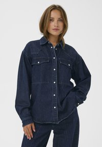 Donkerdenim shirt met een kraag, twee borstzakken en knoopsluiting. Kenmerkt zichtbare stiksels en een ontspannen pasvorm ontwerp.