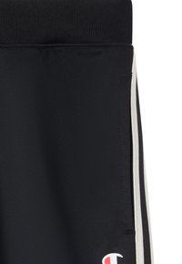 Pantalons de sport noirs avec une taille texturée, des rayures blanches sur les côtés et un logo en bas à gauche. Le matériau semble lisse et extensible.