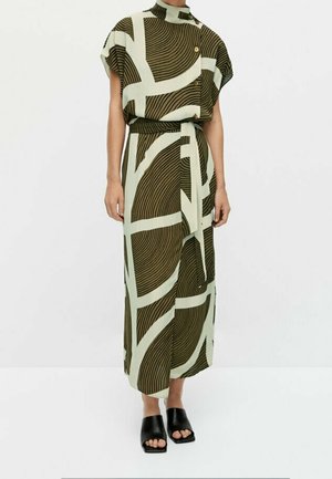 Femme portant une longue robe à motifs avec manches courtes et col montant, associée à des sandales mules noires à bout ouvert.