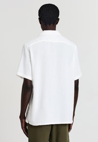 Chemise blanche à manches courtes en tissu texturé, dotée d'un col classique et d'une coupe décontractée, avec des coutures visibles et un ourlet droit.