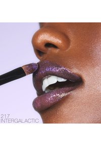 Paarse glanzende lipgloss wordt aangebracht met een kwast, waarbij glinsterende accenten en een glanzende textuur op volle lippen tegen een bruine huidtoon worden benadrukt.