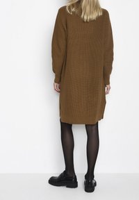 Bruine, lange mouw gebreide jurk; losse pasvorm met zijsplits; gecombineerd met zwarte schoenen. De textuur is ribbelig en chunky.