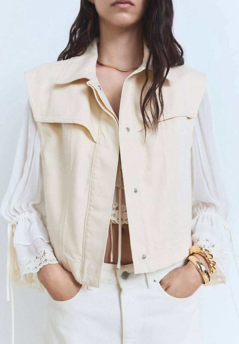 Femme portant une veste sans manches crème sur un chemisier en dentelle blanche à manches bouffantes et un pantalon blanc, mains dans les poches, bracelets en or au poignet.
