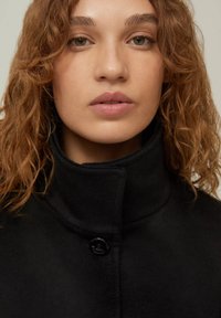 Giacca in pile nera con colletto alto e chiusura a un solo bottone nero. Il design è minimalista e presenta una texture morbida.
