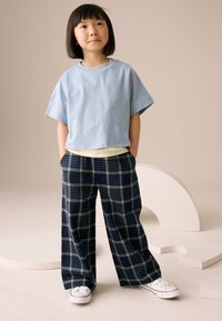 T-shirt azzurro chiaro corta con maniche corte, indossata sopra una maglia beige. Pantaloni a gamba larga a quadri blu navy con dettagli crema e sneakers bianche.