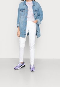 Chaqueta de mezclilla en azul claro con detalles en botones, llevada sobre una camisa de tie-dye en lavanda. Pantalones blancos ajustados y zapatillas Nike moradas completan el look.