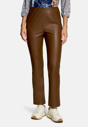 Femme portant un pantalon droit en cuir synthétique marron, associé à des baskets blanches et un haut à manches longues à motifs.