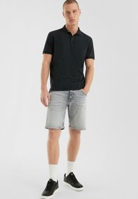 Zwarte polo gecombineerd met lichtgrijze denim shorts. Model draagt zwarte sneakers en witte sokken, staat casual met één hand in zijn zak.