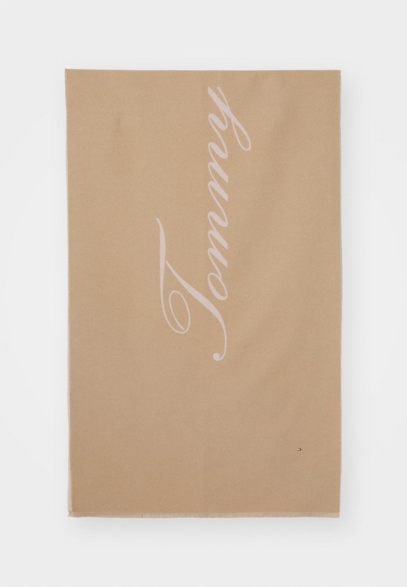 Beige rektangulært stoff med "Tommy" i store kursivbuchstaver og en liten Tommy Hilfiger-logo nær det nederste høyre hjørnet.