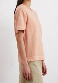 Camiseta de manga corta de color durazno con cuello redondo, hecha de tela suave. Presenta un bolsillo parche en el lado izquierdo del pecho y un dobladillo recto.