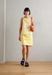 Geel mouwloos topje met gedrapeerd detail, bijpassende gele rok en zwarte sandalen met twee banden; model draagt een kleine rode tas.
