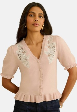 Kobieta ubrana w jasno-różowy kardigan z krótkim rękawem, zapinany na guziki, z kwiatowymi koronkowymi detalami przy ramionach oraz falbaniastym dołem i mankietami.