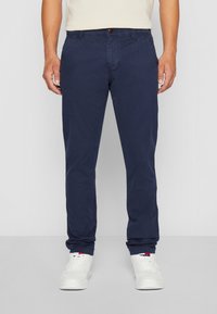 Man som bär marinblå avslappnade byxor och vita sneakers, står mot en lätt enfärgad bakgrund.