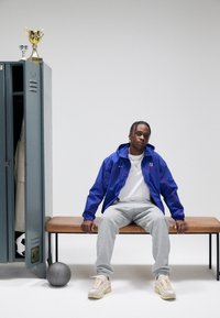 Idrettsutøver i blå jakke med røde detaljer, grå joggebukser og beige joggesko, sitter på en trebenk ved siden av et skap og sportsutstyr.