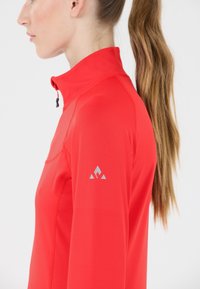 Rode sportieve pullover met een hoge kraag, gemaakt van een gladde, lichte stof. Heeft een klein grijs logo op de rechter schouder.