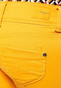 Jeans de mezclilla amarillos con un bolsillo trasero con costuras decorativas y un botón de metal. Etiqueta "Pepe Jeans London" visible en la banda de la cintura.