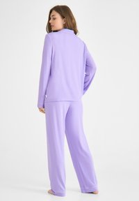 Lila tvådelad loungedräkt i mjukt tyg. Innehåller långärmade tröja, krage och lös passform med rakskurna byxor.