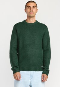 Pull en maille vert foncé avec un col rond, des poignets et un ourlet côtelés, et une finition texturée. Présente un petit logo sur le bas de l'ourlet.